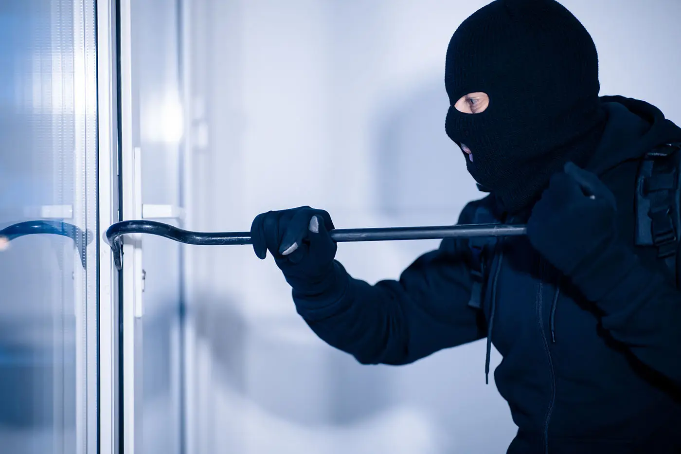 Ladrón encapuchado forzando la puerta de un apartamento con una palanca